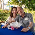 A woman and a man sit closely together at an outdoor table with food and drinks, smiling at the camera. Behind them, the SB Sculpture adds a creative flair to their cheerful gathering. The man wears a name tag that says "Doug.