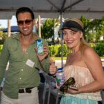 A man and woman stand under a tent at an outdoor SB Sculpture event, each holding a canned drink and smiling at the camera. The man wears sunglasses; the woman wears a visor.