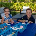 Two children sit at a blue table outdoors, coloring with markers and paper, perhaps inspired by an SB Sculpture. Plastic baskets hold art supplies on the table, with greenery and patio furniture in the background.