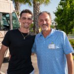 Two men stand outdoors smiling at the camera, with a food truck and palm trees in the background. One man wears a black shirt; the other has a name tag and a blue patterned shirt, near the vibrant SB Sculpture.