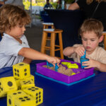 Two young children play with kinetic sand and plastic molds at a table with large yellow foam dice, creating their own SB Sculpture masterpieces. Other people are visible in the background.