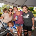 Four adults and a child pose and smile under a tent at an outdoor SB Sculpture event; one woman uses crutches, and all hold drinks.