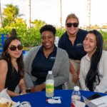 Four women sit at an outdoor table with drinks and food, smiling at the camera. Palm trees, greenery, and a glimpse of the SB Sculpture create a lively backdrop.
