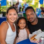 A woman, a man, and a young girl sit closely together at a table outdoors, smiling at the camera with food and drinks in front of them, while the SB Sculpture provides an artistic backdrop to their cheerful gathering.