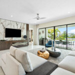 Modern living room with a large sectional sofa, marble accent wall with mounted TV, floor-to-ceiling windows, and a view of a patio and waterway outside.