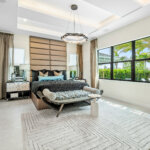 Modern bedroom with large windows, a black bed, accent lighting, neutral walls and floors, a bench at the foot of the bed, and minimalistic decor. Natural light fills the room.