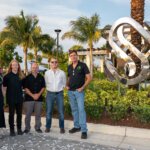 Five people stand outdoors near the SB Sculpture, surrounded by palm trees and landscaping, with a building in the background.