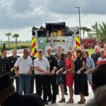 A group of people stands in front of a fire truck holding a ribbon at a ceremony, with fire personnel, palm trees, and cloudy sky in the background.
