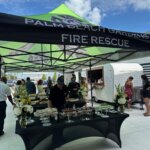 People gather under a Palm Beach Gardens Fire Rescue tent with a table of food and flowers; a mobile cocktail bar trailer is in the background.