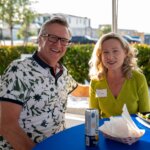 A man and a woman sit at a blue table outdoors, smiling at the camera near the SB Sculpture. They have drinks and food in front of them, and the woman wears a name tag that reads "Cony.