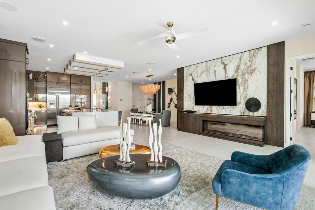Experience Manhattan elegance in this modern open-plan living room featuring a blue armchair, white sofas, metallic coffee table, large TV above a marble fireplace, and sweeping views into the Solana Bay kitchen and dining area.