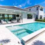 Modern backyard with a rectangular swimming pool and attached hot tub, surrounded by a tiled patio and outdoor seating area, next to a white house under a clear blue sky.