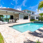 Modern house with a covered patio, outdoor dining area, swimming pool, water feature, and landscaped backyard on a sunny day.