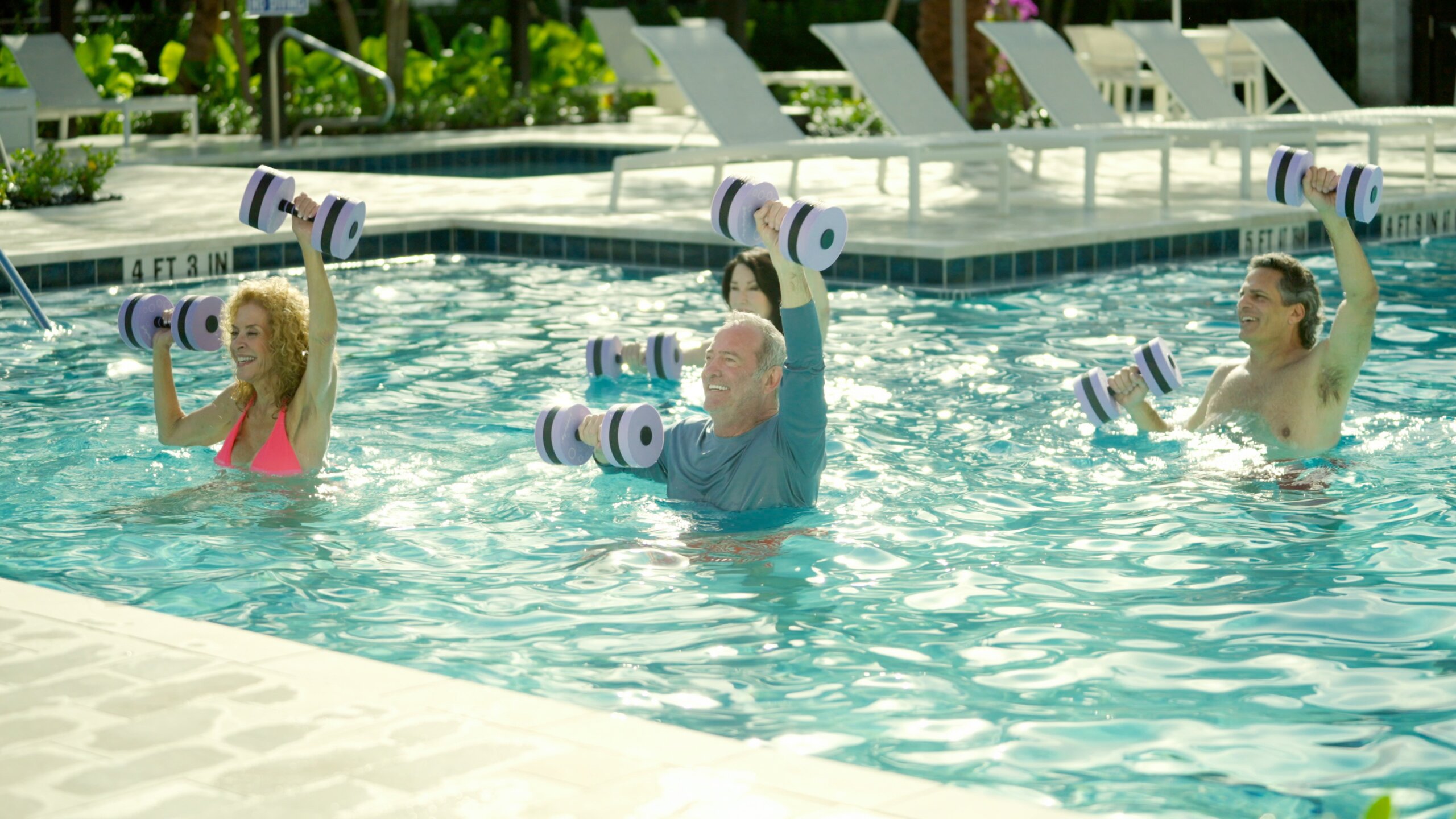 Four adults in a swimming pool participate in a water aerobics class, lifting foam dumbbells above their heads near the pool edge, enjoying an energizing Catalina Palms experience.